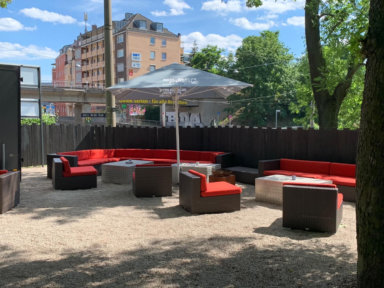 Lounge-Ecke im Biergarten mit roten Polstern auf Kies, dazu ein Sonnenschirm und Bäume