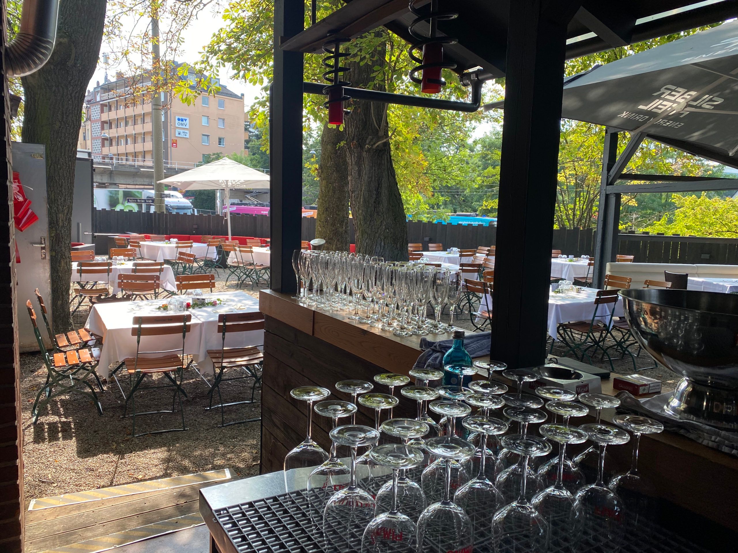 Gläser auf der Außenbar, dahinter der Biergarten mit gedeckten Tischen unter Bäumen