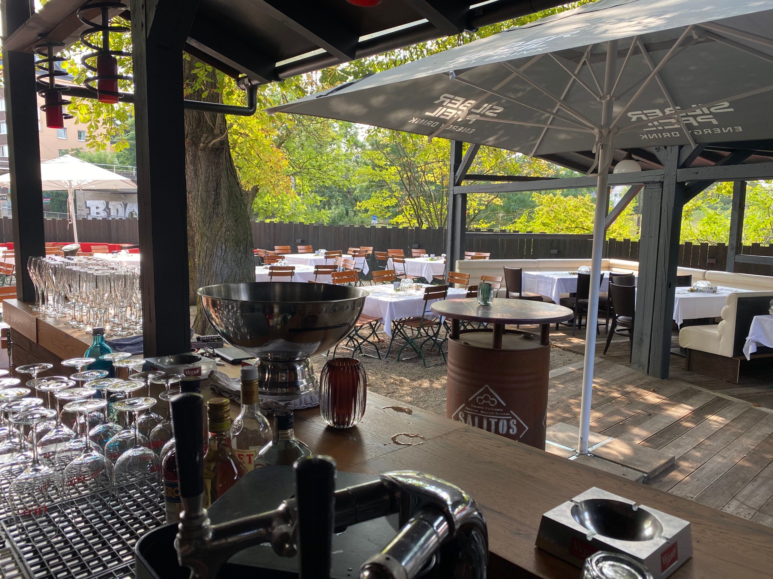 Blick von der Außenbar unter dem Holzdach in den Biergarten mit Kies, großen Bäumen und gedeckten Tischen