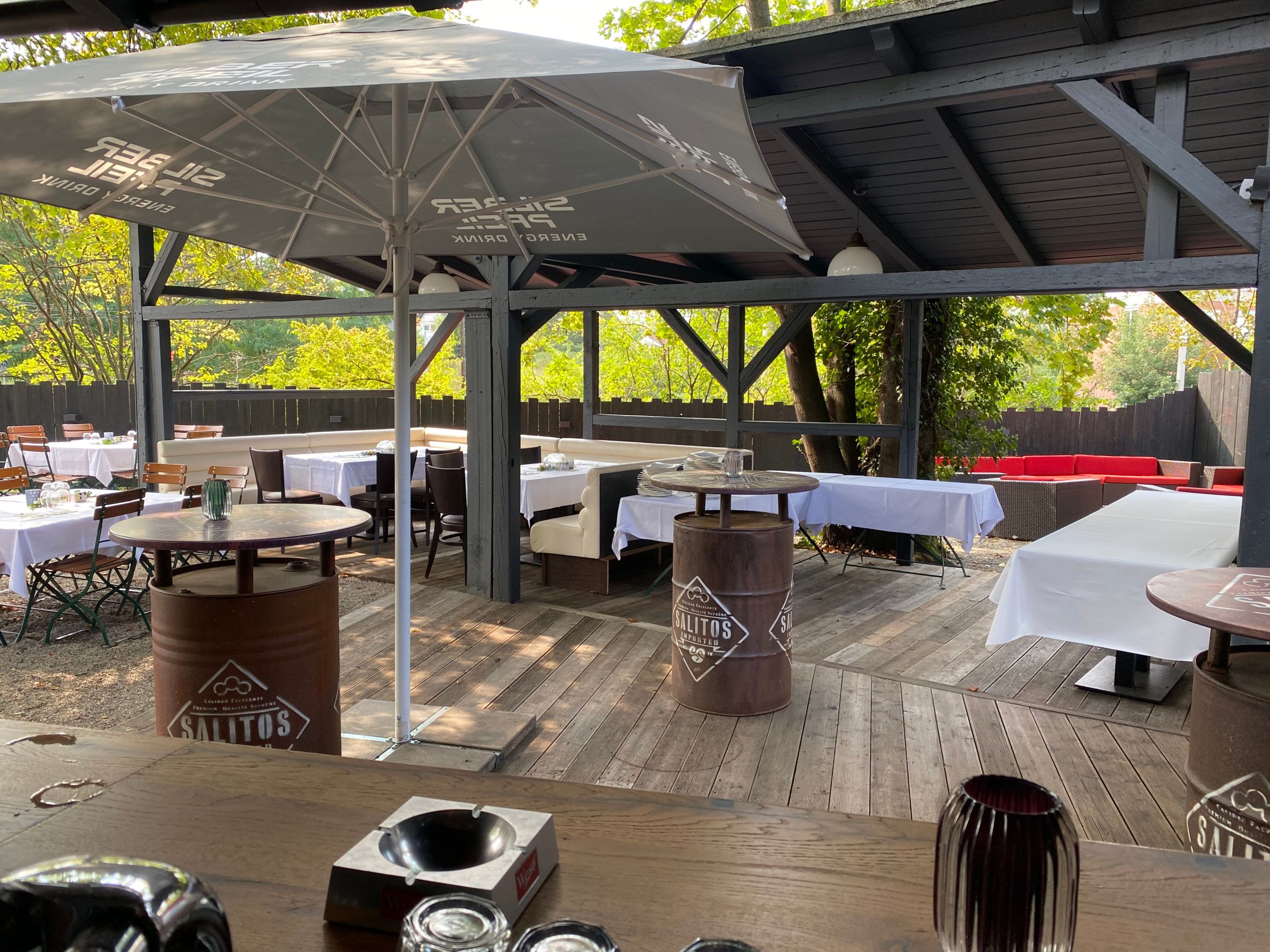 Überdachte Holzterrasse im Biergarten mit Stehtischen aus Fässern, Sonnenschirm und gedeckten Tischen