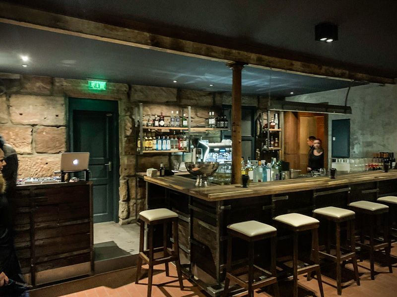 Bar mit Holztheke und hellen Barhockern vor der Natursteinwand, links ein DJ-Pult